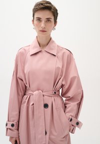 Roza trench kaput s pojasom u struku, velikim reverima i oversized rukavima. Ima crne gumbe i bočne džepove za dodatne detalje.