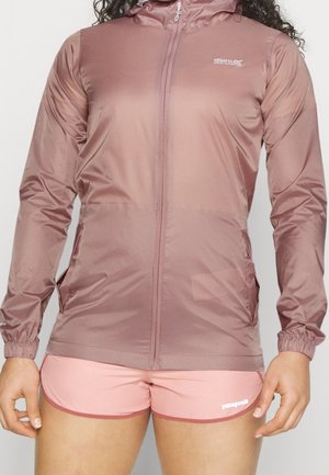 Vrouw draagt een mauve windbreaker jas met ritssluiting en capuchon en roze sportshorts tegen een neutrale achtergrond.