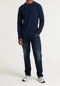 Marineblauer strukturierter Strickpullover mit Rundhalsausschnitt, kombiniert mit dunklen Jeans und hellgrauen Sportschuhen mit silbernen Akzenten.