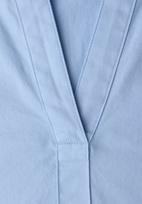 Tissu bleu clair à texture lisse présentant un col en V. Les coutures visibles soulignent les angles nets du col.