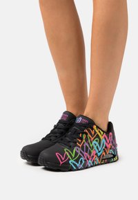Schwarze Sportschuhe mit bunten Herzmustern in Pink, Orange, Grün und Blau. Mit einem Schnürdesign und gepolsterten Sohlen.