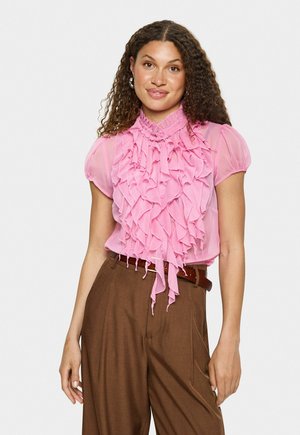 Blouse rose clair avec des détails froncés sur le devant, manches courtes bouffantes et col montant. Associée à un pantalon brun taille haute et une ceinture.