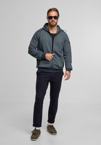 Blaue Kapuzenjacke aus strukturiertem Stoff, mit Reißverschluss und verstellbarem Kapuzen. Kombination mit dunklen Hosen und braunen Schnürschuhen.