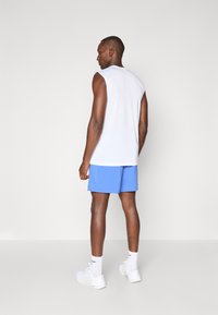 Hvid ærmeløs skjorte og blå shorts. Skjorten er glat og tætsiddende; shortsene er lette. Modellen bærer hvide sportssko.