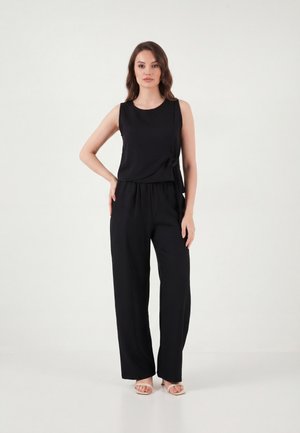 Femme debout, une main sur la hanche, portant un haut noir sans manches avec un nœud sur le côté et un pantalon noir ample, sur fond blanc.