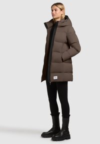 Brauner Puffercoat mit Kapuze und schwarzem Reißverschluss, der leicht oversized geschnitten ist und eine gesteppten Textur aufweist. Kombiniert mit schwarzen Hosen und Stiefeln.