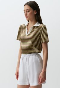 Polo a maniche corte con motivo a righe in tonalità verdi, marroni e gialle, con colletto bianco; abbinato a pantaloni corti bianchi con laccio.