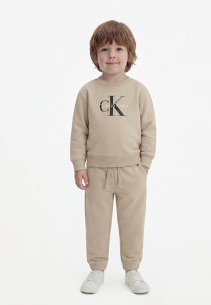 Małe dziecko stojące, ubrane w beżową bluzę Calvin Klein, pasujące spodnie dresowe i białe trampki na jednolitym tle.