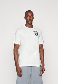 Camiseta blanca de manga corta con un logotipo y un escudo negro de "Raiders" en el lado izquierdo del pecho, combinada con pantalones cortos grises. Textura de tela suave.