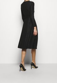 Nainen, joka on pukeutunut pitkähihaiseen mustaan midi-mekkoon, jossa on istuva vyötärö, ja mustiin korkokenkiin, seisoo valkoista taustaa vasten.