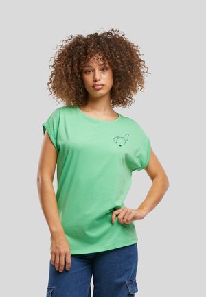 Femme aux cheveux bouclés portant une chemise vert clair aux manches retroussées avec un petit motif de visage de chien et un pantalon cargo bleu, debout devant un fond gris.