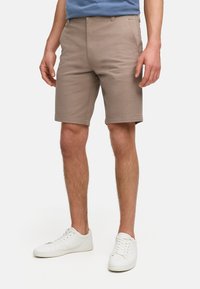 Shorts en coton marron clair avec un devant plat, des passants de ceinture et des poches latérales. Associés à des baskets blanches sur un fond neutre.