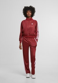 Burgundský sportovní teplákový komplet s texturovanou tkaninou, bílé pruhy na kalhotách a rukávech, bunda na zip a boční kapsy. Bílé tenisky.