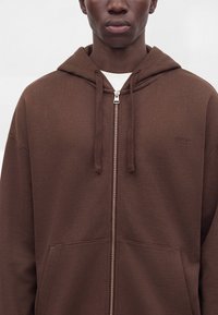 Homme portant un sweat à capuche marron zippé avec capuche et cordons, arborant un marquage subtil "HUGO" sur la poitrine.