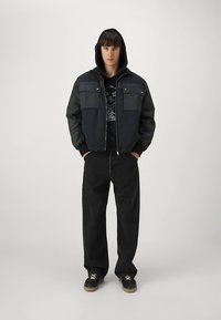 Casaco preto com capuz, mangas contrastantes, fecho de correr e dois bolsos ao peito. Combinado com calças pretas de corte largo e ténis pretos.