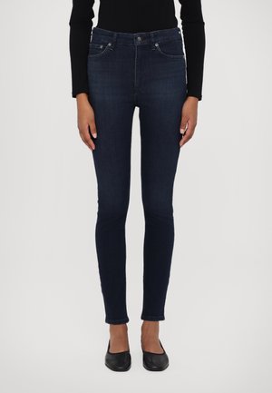 Femme portant un jean skinny bleu foncé, un haut à manches longues noir et des chaussures plates noires, debout devant un fond clair uni.
