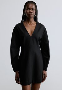Μαύρο φόρεμα με βαθύ V λαιμόκοψη και μακριά μανίκια. Διαθέτει δομημένους ώμους και εφαρμοστή σιλουέτα. Κατασκευασμένο από λείο, υφασμένο ύφασμα.