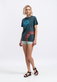 Korte mouwen donkergroen T-shirt met een blauwe cirkelvormige afbeelding en een rood abstract ontwerp, gecombineerd met lichtmintkleurige shorts en zwarte sandalen.