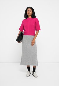 Pull en tricot fuchsia avec un motif en relief, associé à une jupe maxi côtelée grise. Le mannequin tient un sac à main noir, porte des chaussures blanches et des chaussettes noires.
