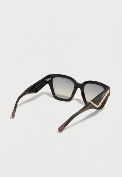 Μαύρα γυαλιά ηλίου σχήματος μάτι γάτας με φακούς βαθμιαίας μετάβασης και χρυσές λεπτομέρειες στους βραχίονες, εμφανιζόμενα από πίσω σε λευκή επιφάνεια.