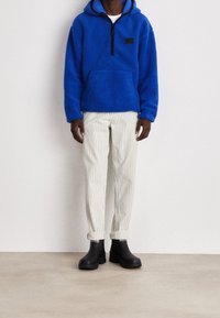 Felpa blu in pile con mezza zip e tasca frontale, abbinata a pantaloni bianchi a righe e stivali neri alla caviglia. Sfondo bianco.