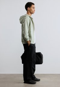 Νεαρός άνδρας στέκεται σε προφίλ φορώντας ανοιχτοπράσινο φούτερ με φερμουάρ, μαύρο παντελόνι και παπούτσια, κρατώντας μια μαύρη τσάντα duffel μπροστά σε μονόχρωμο φόντο.