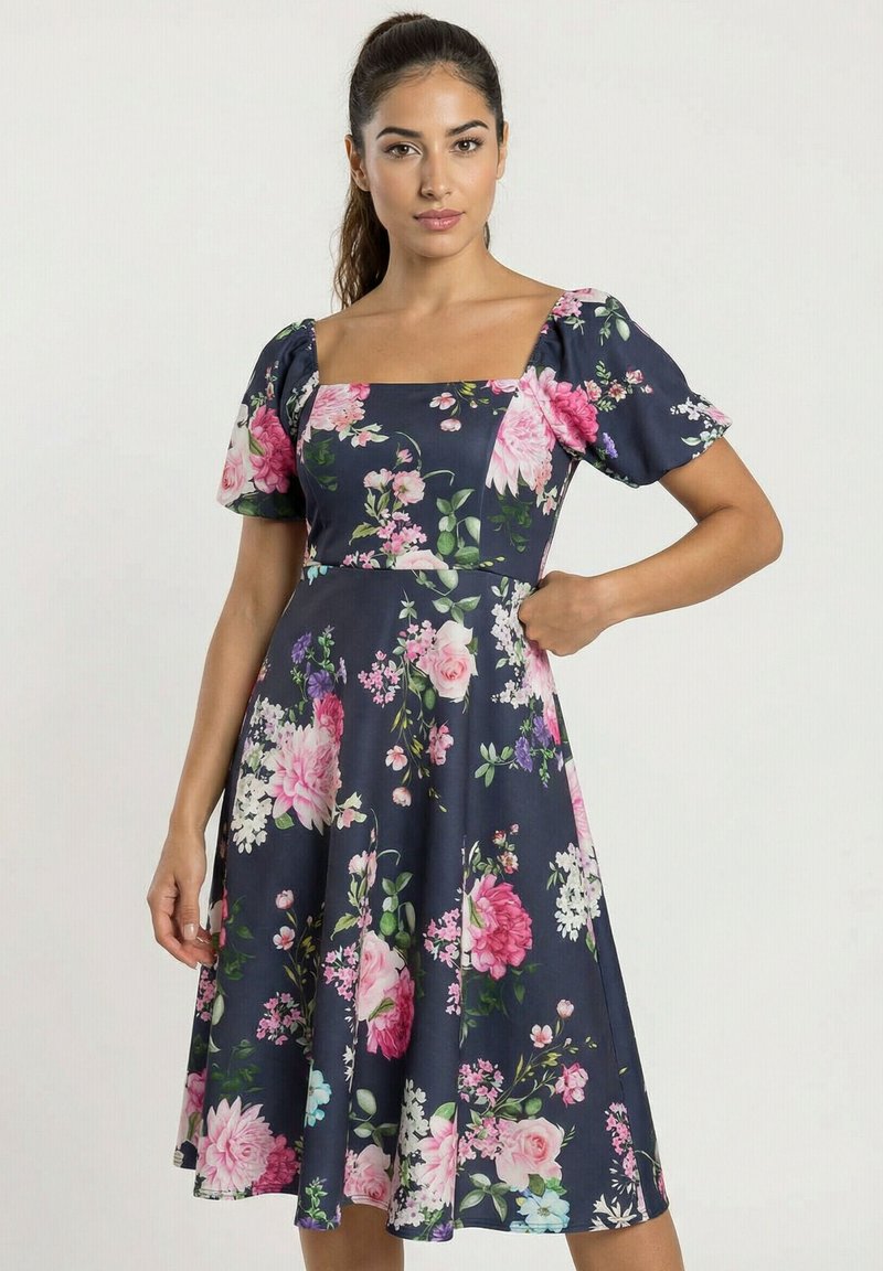 Frau in einem marineblauen knielangen Kleid mit rosa-weißem Blumenmuster, Puffärmeln und quadratischem Ausschnitt, posiert vor einem schlichten Hintergrund.