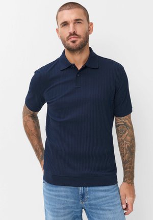 Man met kort haar en baard, draagt een donkerblauw polo met korte mouwen en lichtblauwe spijkerbroek, handen rustend bij de zakken, effen achtergrond.