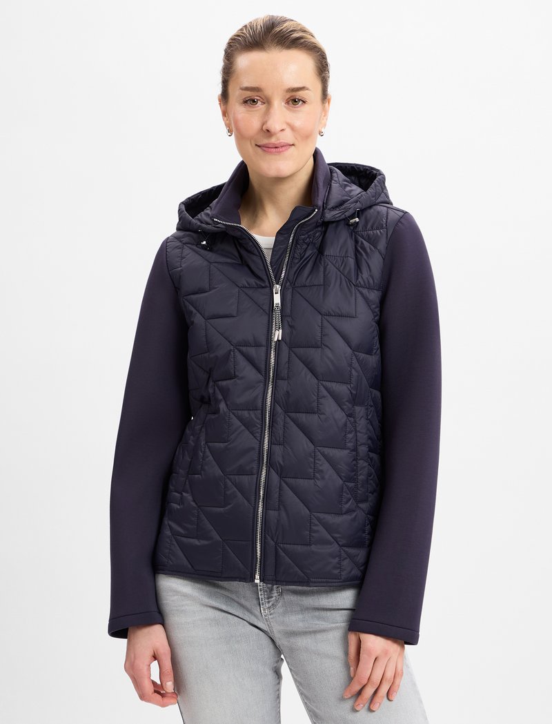 Marineblauer gesteppter Weste mit Kapuze, mit Reißverschluss vorne, hohem Kragen und kontrastierenden Strickärmeln. Strukturmuster mit silbernen Hardware-Akzenten.