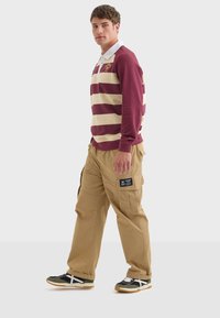 Maroone og creme stribet langærmet polo skjorte parret med tan cargo bukser. Cargo bukserne har sidelommer og et logo patch. Sorte og grønne sneakers.