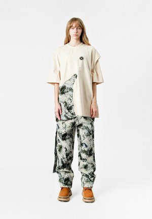 Joven mujer con largo cabello rubio viste una camiseta oversized beige con un patrón abstracto y pantalones a juego, de pie frente a un fondo blanco.