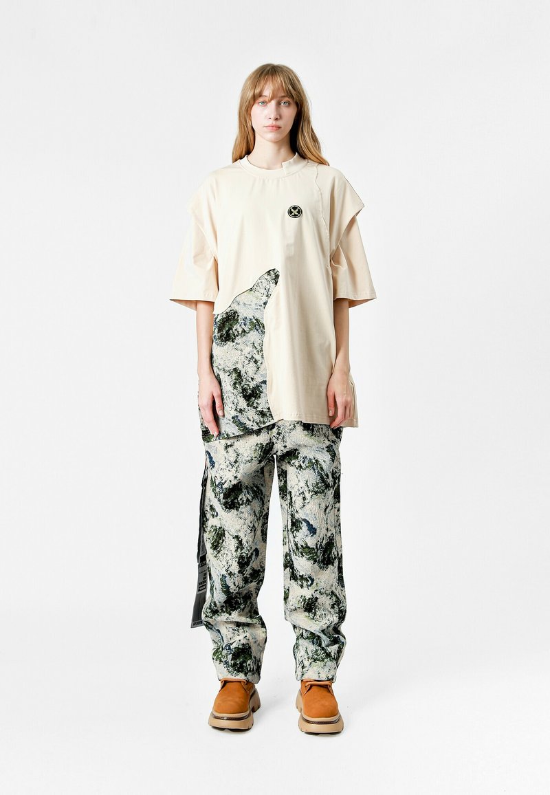 Joven mujer con largo cabello rubio viste una camiseta oversized beige con un patrón abstracto y pantalones a juego, de pie frente a un fondo blanco.