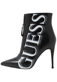 Botín negro de cuero hasta el tobillo con puntera puntiaguda y tacón de aguja. Presenta un gran logo "GUESS" en gris en el lateral y un detalle de cremallera plateada.