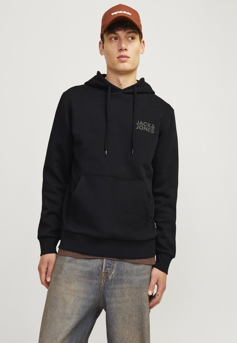 Jeune homme portant un sweat à capuche noir Jack & Jones, un jean bleu et une casquette marron, regardant sur le côté sur un fond uni.