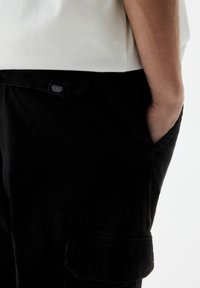 Zwarte cargobroek met een gladde textuur, voorzien van zijzakken en een label met de tekst "UPDATED PANTS" op de achterkant.