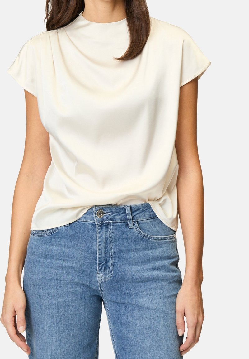 Femme portant un chemisier blanc à manches courtes rentré dans un jean bleu taille haute, avec de longs cheveux bruns partiellement visibles.