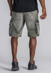 Pantalones cortos de carga en denim verde oliva deslavado con múltiples bolsillos laterales y costuras visibles. Llevado con zapatillas negras y una camiseta negra.