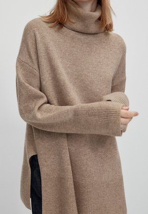 Kvinde iført oversized beige rullekrave sweater med lange ribbede manchetter og sideslids, der holder i den ene manchet med hånden.