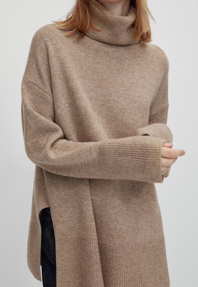 Femme portant un pull à col roulé beige oversize avec de longs poignets côtelés et une fente latérale, tenant un poignet dans sa main.