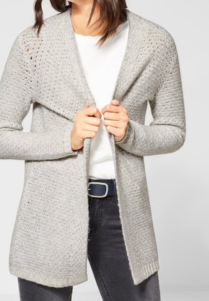 Woman wearing textured light gray open-front cardigan over white shirt and dark jeans, holding edges of the cardigan at chest.