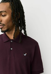 Tricou polo burgundy cu material texturat, având un placket cu trei nasturi și un mic logo brodat cu pasăre pe pieptul stâng.