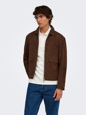Jeune homme portant une veste en daim brun foncé par-dessus un polo blanc et un jean bleu, debout avec les mains détendues le long du corps, regardant légèrement à gauche.