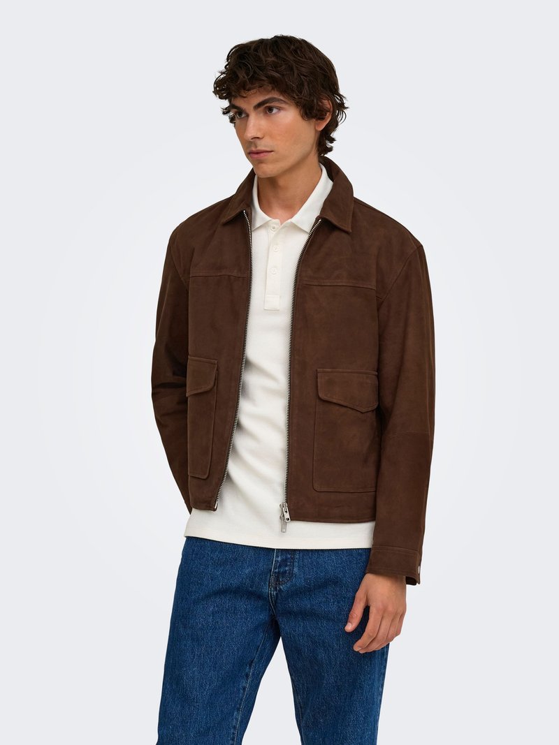 Jeune homme portant une veste en daim brun foncé par-dessus un polo blanc et un jean bleu, debout avec les mains détendues le long du corps, regardant légèrement à gauche.