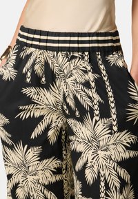 Zwarte stoffen broek met een palmbladerenpatroon in crème. Elastische tailleband met gestreepte details. Zijdelijjke zakken zichtbaar.