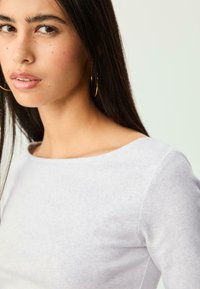 Light gray, long-sleeve top with a rounded neckline, soft texture, and visible seam detailing; model wearing gold hoop earrings.