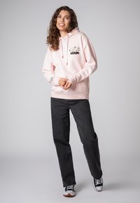Lichtroze hoodie met een voorzak, rode koorden en een grafisch ontwerp. Gecombineerd met zwarte broek en zwart-witte sneakers.