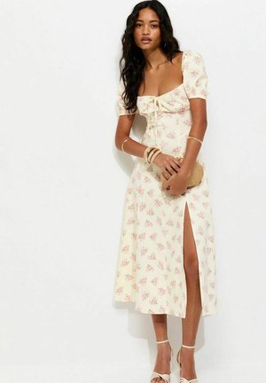 Femme portant une robe midi crème à motifs floraux avec manches bouffantes et fente avant, tenant une pochette tissée et portant des talons beiges et des bracelets.