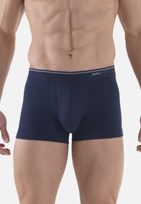 Torso maschile e parte superiore delle gambe che indossa boxer blu navy con una cintura brandizzata su uno sfondo bianco semplice.