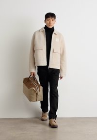 Beige fleecejacka med två framfickor, svart polotröja, svarta byxor och bruna skor. Bär en brun och beige duffelväska.
