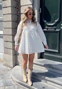 Abito bianco in pizzo con maniche a palloncino lunghe, scollatura con cordoncino e silhouette svasata, abbinato a stivali da cowboy ricamati beige.